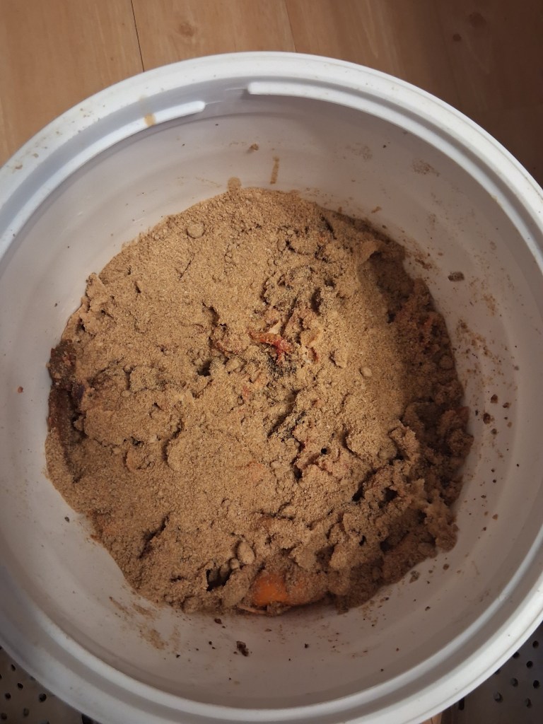 Top-down view of Bokashi bran layered and pressed over fresh kitchen fruit scraps inside a white 5-gallon bucket for anaerobic fermentation