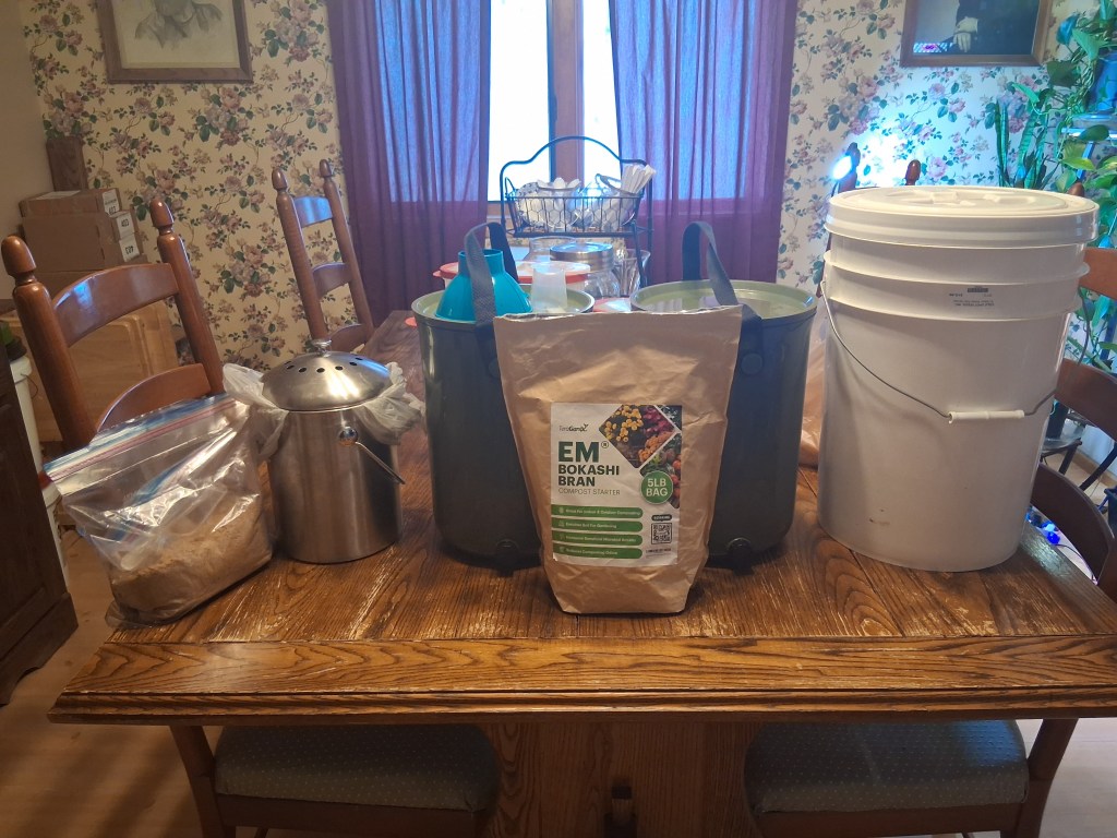 Indoor Bokashi composting setup on wooden table in Alaska home showing EM Bokashi bran bag, green fermenting buckets with spouts, stainless steel kitchen collector, and white 5-gallon bucket